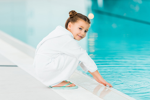 cheerful kid in bathrobe touching water in swimming pool