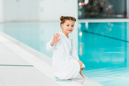 niño lindo sentado en la piscina de la bata de baño y saludando la mano diciendo Hola photo