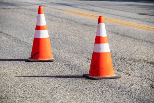 Two grungy orange traffic cones with reflective tape and sharp shadows on cracked asphalt pavement with double yellow line - close-up and selective focus stock photo
