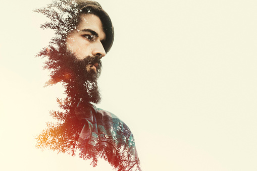 A profile portrait of a bearded man, double exposed in camera so that his image is composited with a line of trees, the sun casting glowing light from behind.  Horizontal with copy space.