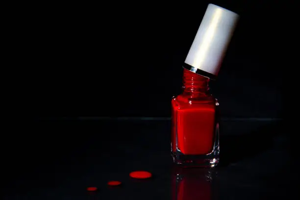 Open bottle with red nail polish. Brush touching the bottle. Drops on the table. Black background.