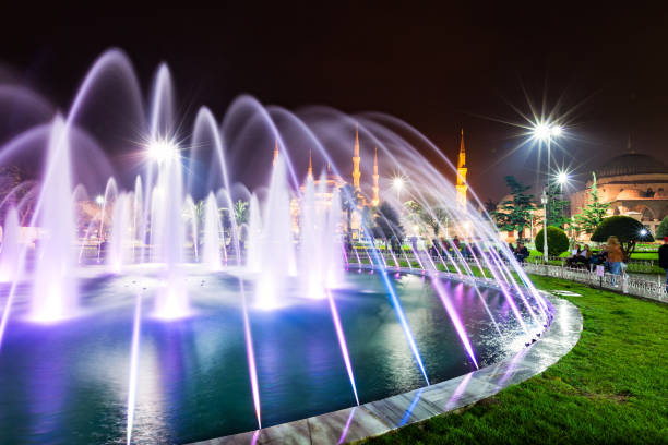 People sit besides an illuminated fountain in Sultanahmet Park, with the Blue Mosque in the background.