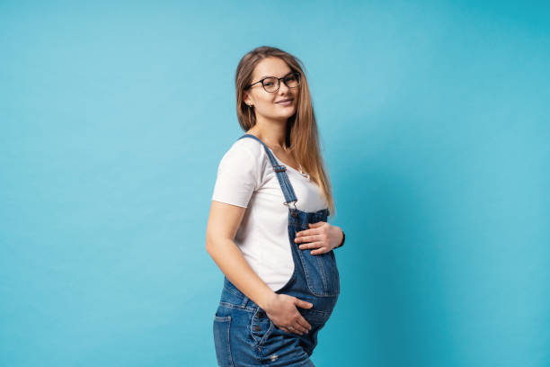 Smiling pregnant woman wearing glasses caressing her belly over blue background stock photo