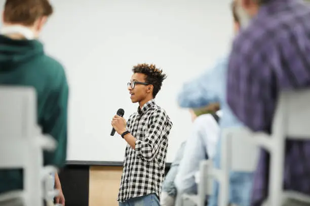 Cheerful confident young black student with Afro hairstyle standing in convention center room and presenting his startup project at conference