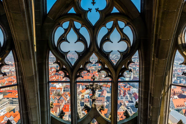 Dramatic, vertical view of a a small leaded window seen atop a medieval, European cathedral. The thick stone walls can be seen.