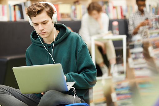 Serious hipster student boy listening to music in headphones and using laptop while searching for information on Internet, student in library concept