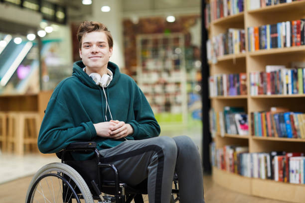 Content disabled student in library Content handsome young disabled student with headphones on neck siting in wheelchair and looking at camera in modern library or bookstore library photos stock pictures, royalty-free photos & images