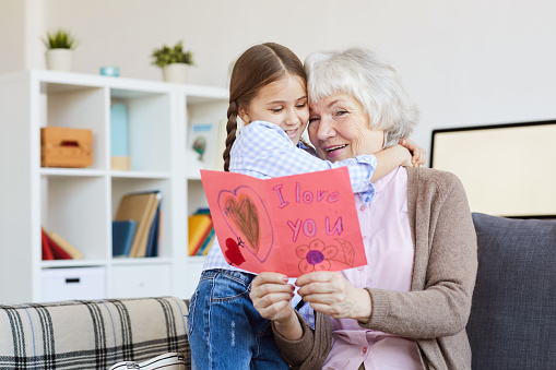 Portrait of cute little girl hugging grandma and giving her handmade I love you card on Valentines day, copy space