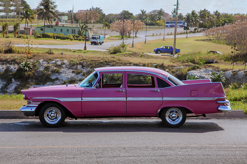 Classic bright pink vintage / retro car of 1950s from side proection, settled in front of a village landscape. Outdoor, copy space.