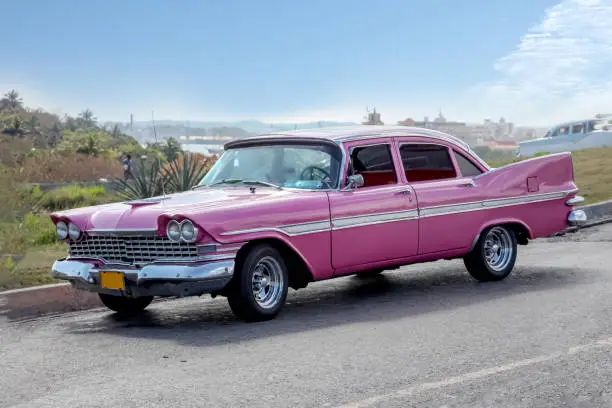 Photo of Classic bright pink vintage / retro car of 1950s from side, settled in front of a village landscape.