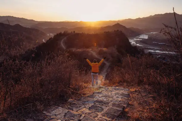 Photo of Woman hiking on mountain path from rear view raises arms to celebrate with feeling good-relax-freedom at sunset-sunrise above mountains- traveling adventure-lifestyle-outdoor activity-nature adventure