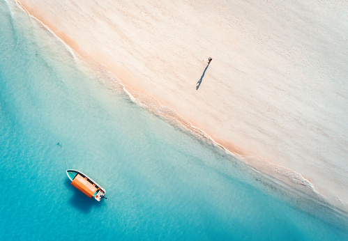 Aerial view of the fishing boat in clear blue water and man on the sandy beach at sunset in summer. Top view of boat. Indian ocean. Travel in Zanzibar, Africa. Landscape with motorboat, sea. Seascape