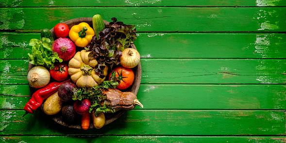 Fresh vibrant colorful organic vegetables in an antique round wooden bowl on an old green wood paneled table background,  atmospheric rustic mood, with good copy space at the right of the image. Vegetables include lettuce, pumpkin, carrot, beetroot, chili, capsicum, onion, tomato, cucumber, avocado and sweet potato.