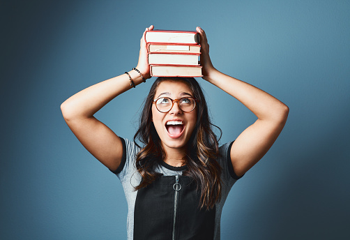 Studio shot of an attractive young woman balancing a pile of books on her head against a blue background