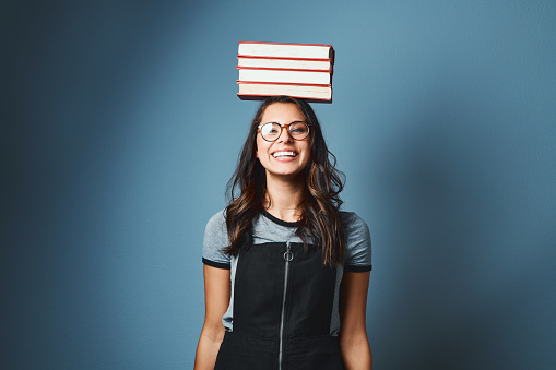 Studio portrait of an attractive young woman balancing a pile of books on her head against a blue background