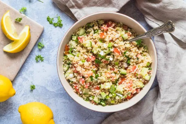 A large bowl of fresh organic healthy Tabouli tabboleh salad with bulgur, tomato, cucumber, lemon and parsley. There are some lemons next to the bowl. Seen from above flat lay perspective.