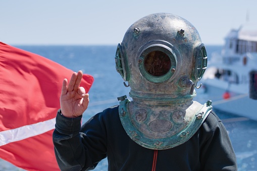 Diver wearing historical metallic helmet showing signal OK, diving flag and boat on the background, historical diving equipment