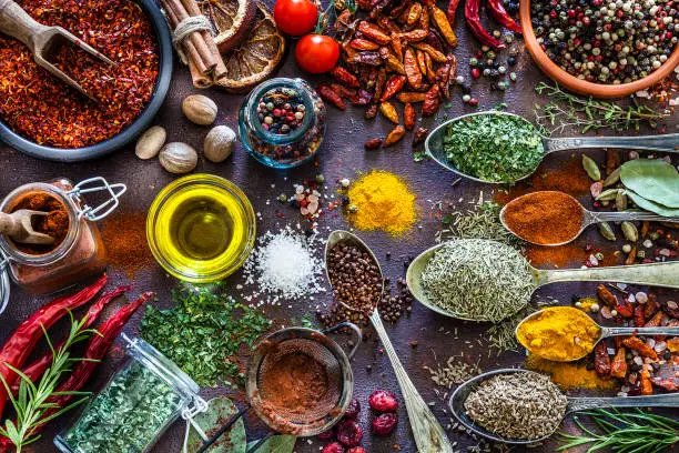 Top view of a rustic brown table filled with a large group of multi colored spices and herbs placed directly on the table. Spices and herb included are clove, turmeric, bay leaf, cinnamon, curry powder, nutmeg, peppercorns, salt, chili pepper, cardamom, dried oregano, parsley, rosemary and dried orange slices. An olive oil bowl is included in the composition. DSRL studio photo taken with Canon EOS 5D Mk II and Canon EF 70-200mm f/2.8L IS II USM Telephoto Zoom Lens