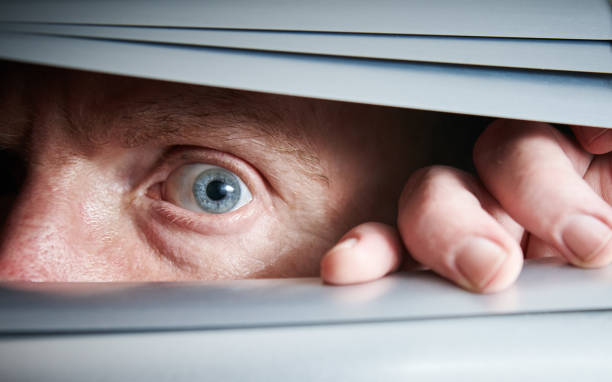 istock Man's single blue eye peeps fearfully through closed venetian blinds 1141622849