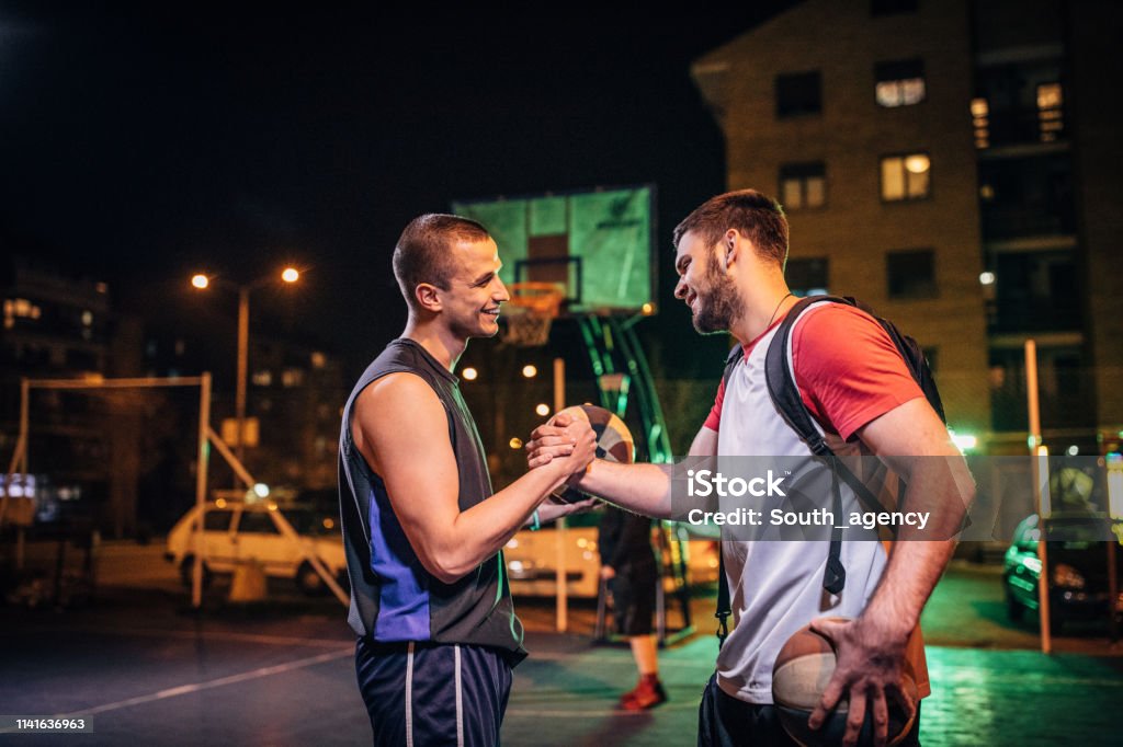 Streetball friends handshake after game Group of men, streetball players standing on court at night downtown in city, handshake after the game. Basketball - Sport Stock Photo