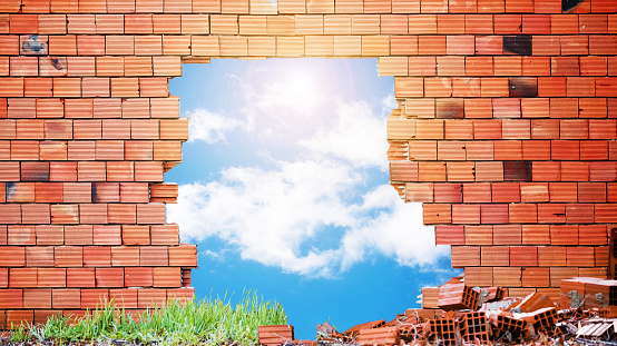 Broken red  brick wall with a big hole in the middle and blue sky, clouds and sun view through the hole.
