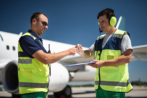 Teamwork at airdrome. Man in sunglasses taking pen and documents for signing while colleague holding clipboard. Blue sky and passenger plane on blurred background