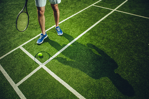 Close up male feet with shadow situating on green field. He keeping racket while playing tennis