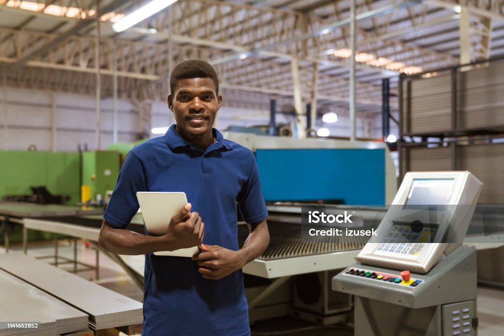 Confident trainee holding digital tablet in industry Portrait of confident trainee holding digital tablet. Male is smiling while standing in industry. He is in uniform at factory. Engineer Stock Photo