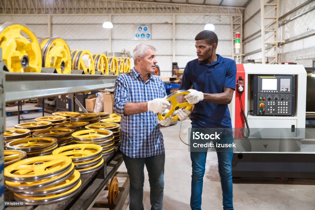 Instructor and trainee holding wheel in factory Male instructor and trainee holding yellow wheel. Engineer is with apprentice in industry. They are standing in factory. Industry Stock Photo