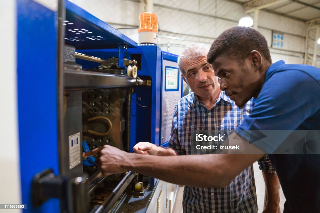 Instructor discussing with trainee over machinery Mature instructor guiding apprentice in using injector machine. Engineer teaching male trainee in industry. They are working in factory. Engineer Stock Photo