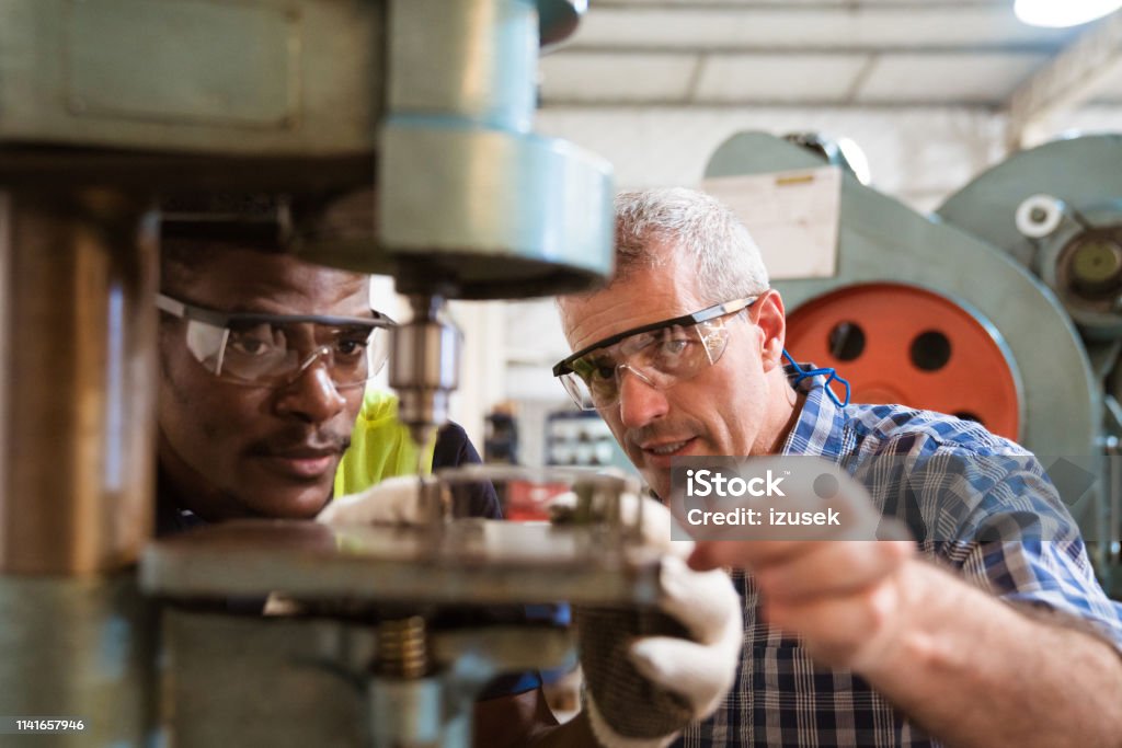 Instructor guiding trainee in using yoke machine Mature instructor guiding trainee in using yoke machine. Engineer is teaching male apprentice in factory. They are working in industry. Factory Stock Photo