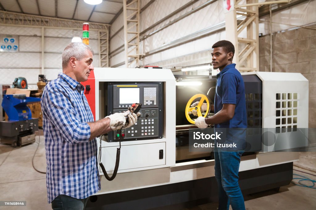 Instructor teaching apprentice in factory Mature instructor teaching apprentice in factory. Male trainee holding wheel is while engineer using remote control. They are working in industry. African Ethnicity Stock Photo