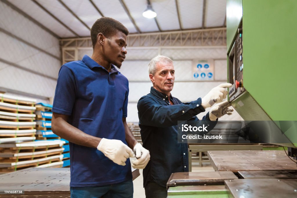 Instructor teaching male trainee in factory Mature engineer explaining apprentice over manufacturing machinery. Instructor is discussing with trainee in factory. They are standing in industry. Industry Stock Photo
