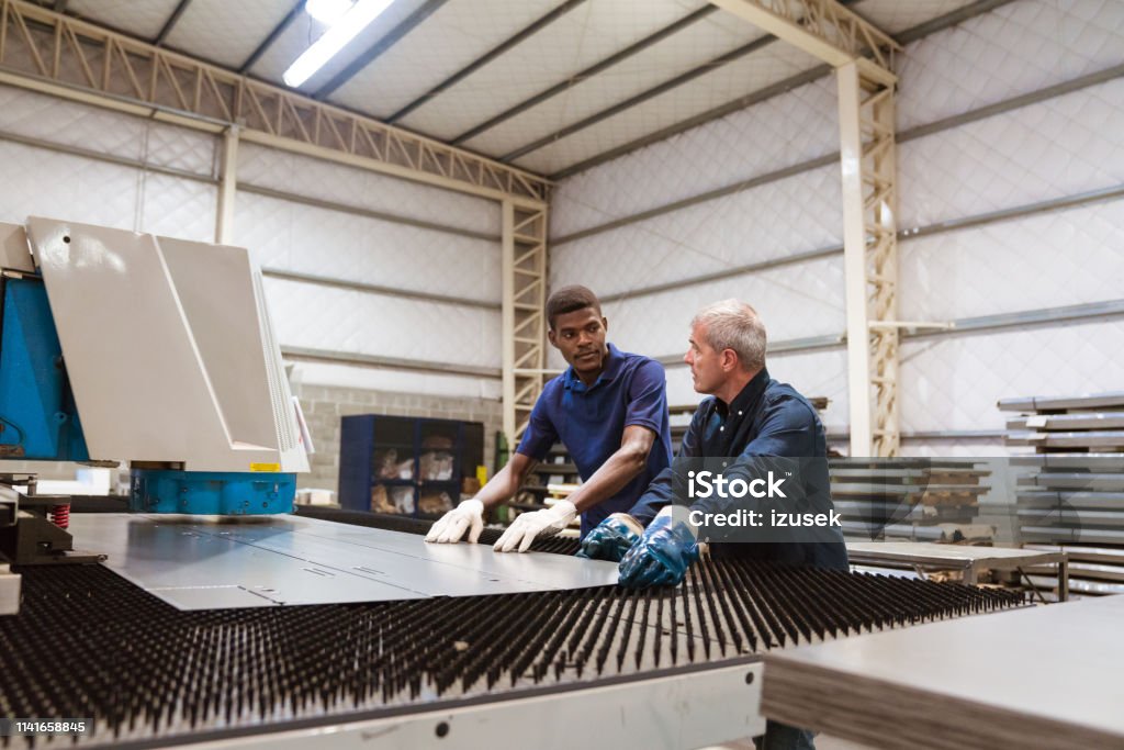 Instructor teaching trainee in factory Mature engineer teaching apprentice in factory. Instructor is discussing with trainee over puller machine. They are working in industry. African Ethnicity Stock Photo