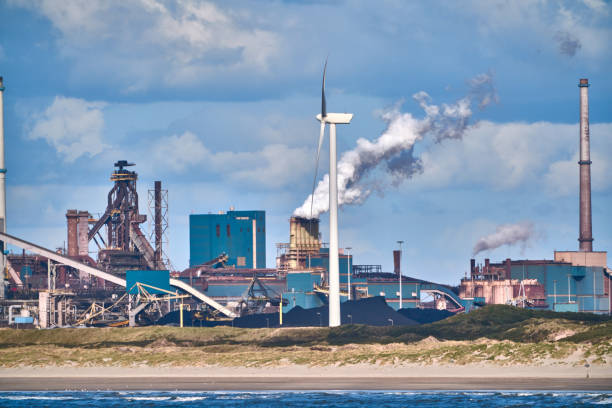 A single wind turbine stands on the edge of an old rusty industrial plant with steaming chimney. As synonymous for the green and eco-friendly evolution instead of a coal-fired power station.