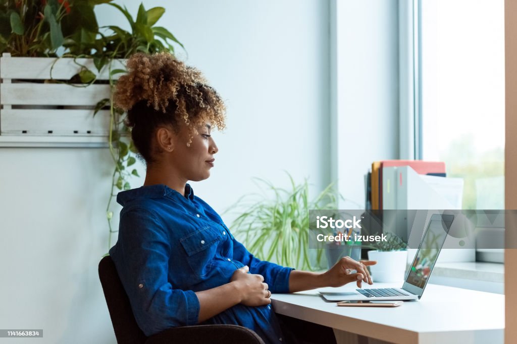 Pregnant woman with hand on belly using laptop Pregnant businesswoman with hand on stomach working at desk. Female professional is wearing business casuals. She is using laptop in office. Pregnant Stock Photo
