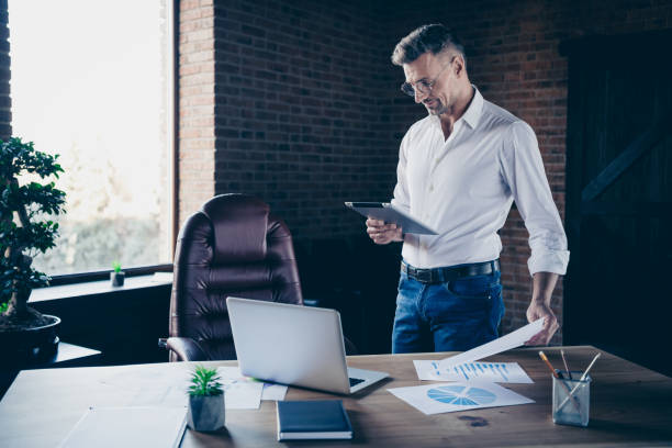 istock Close side profile up photo handsome he him his macho guy ponder pensive attentive look e-book e-reader watch webinar check numbers notebook table stand office wear specs formal wear white shirt 1141664037