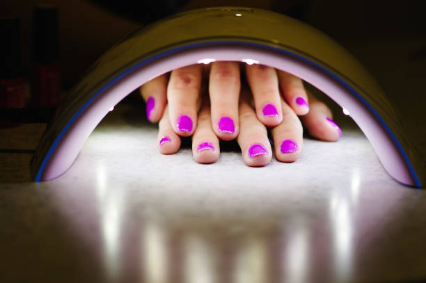 dos mujeres de mano dentro de la lámpara para clavos en la mesa de cerca. lámpara uv para secar clavos con método de gel. uñas violetas secas en la lámpara - fingernail acrylic women beauty fotografías e imágenes de stock