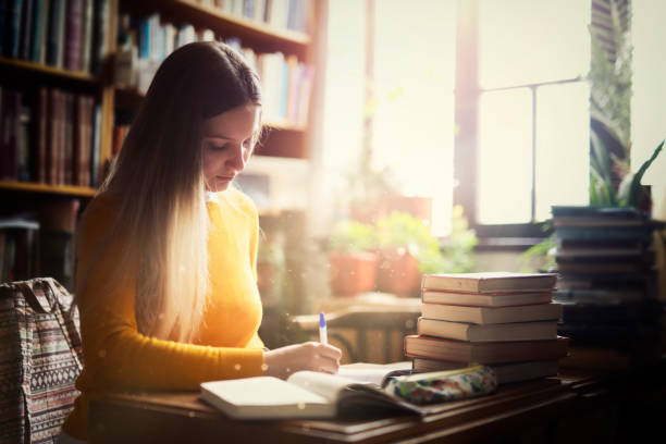 istock Female student using books for taking notes to study, young women in library at dramatic light 1141685558