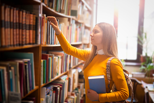 Young female student crouching by the lower shelves and looking for a book in university library