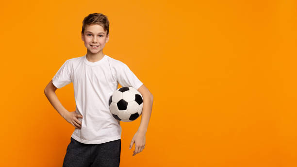 boy posando con balón de fútbol en fondo de estudio naranja - t shirt child white portrait fotografías e imágenes de stock