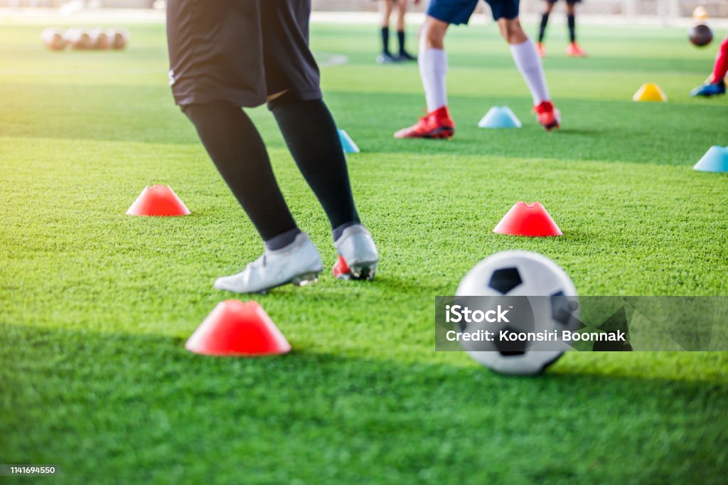 selective focus to red marker cones with blurry soccer ball and kid soccer player Jogging and jump between cone markers selective focus to red marker cones with blurry soccer ball and kid soccer player Jogging and jump between cone markers on green artificial turf for soccer training. Football or Soccer Academy. Soccer Stock Photo