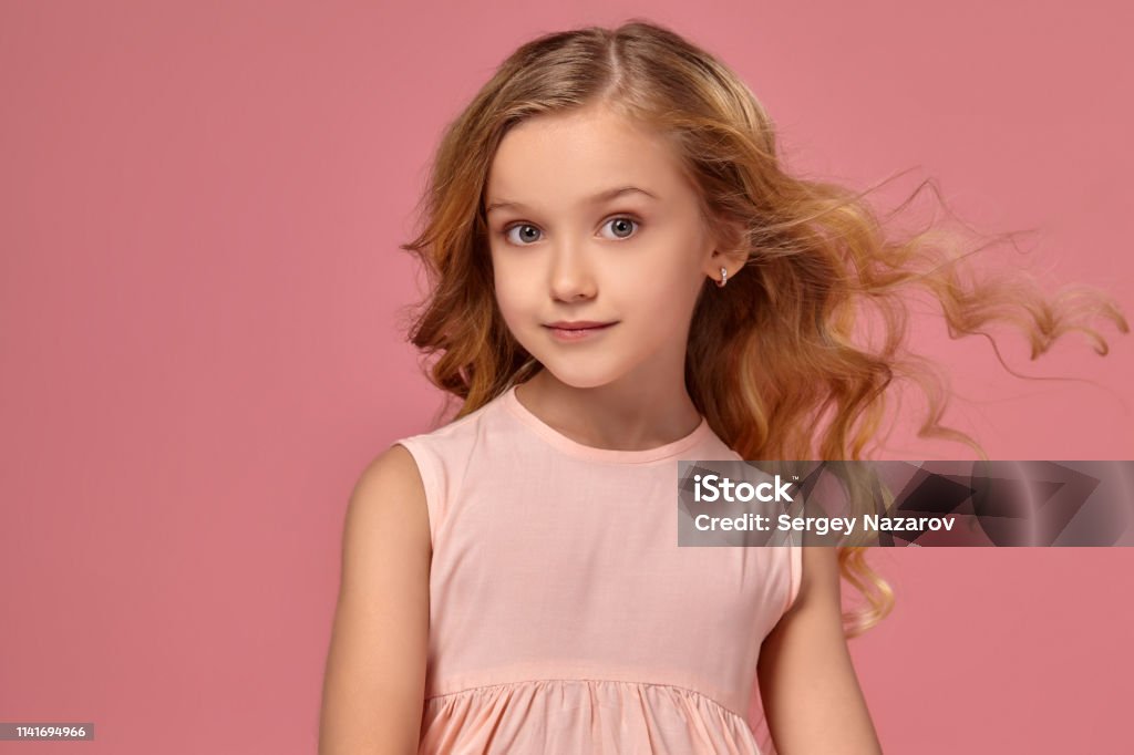 Little girl with a blond curly hair, in a pink dress is posing for the camera Beautiful little girl with a blond curly hair, in a pink dress is posing for the camera and her hair is fluttering, on a pink background Blond Hair Stock Photo