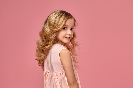 Beautiful little girl with a blond curly hair, in a pink dress is standing sideways and smiling, on a pink background