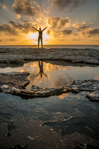 Woman (girl) with outstretched hands situated on rock between sea and puddle. Sunset. Dusk.