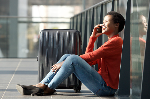 Side portrait of happy young black woman sitting on floor with luggage and talking on mobile phone in airport terminal