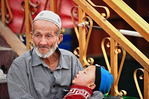 Hotan, China-October 3, 2017: Blue-eyed grandfather of the Uyghur Turkic people holds his napping grandson in his arms while they both sit on the steps of a store at the city's bazaar. Xinjiang Region