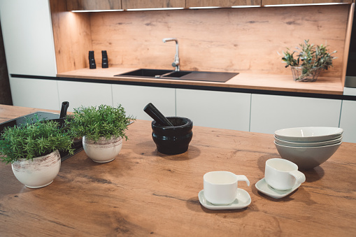 An empty kitchen in white and natural wood combination with black electric appliances, white dining set and fresh herbs.