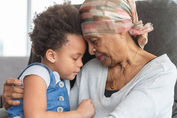 Senior woman with cancer lovingly holds granddaughter A black woman with cancer is wearing a scarf on her head. She is sitting in a lounge chair with her young granddaughter. The two are embracing and their foreheads are touching. They have somber and thoughtful expressions. survival stock pictures, royalty-free photos & images