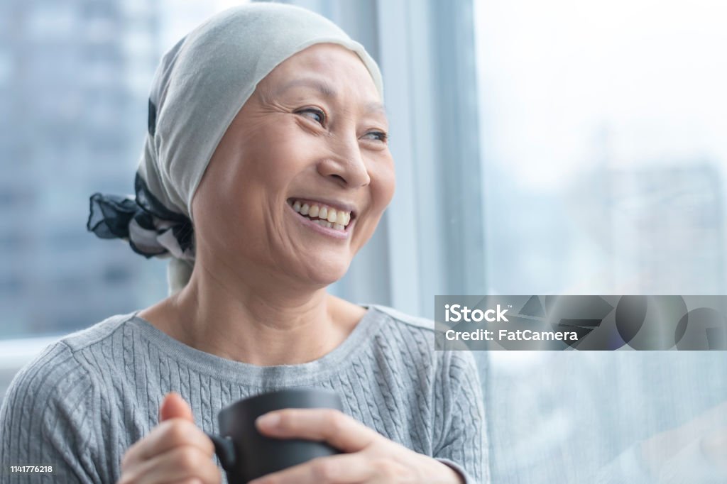 Beautiful Korean woman with cancer looks out window A Korean senior woman with cancer is wearing a scarf on her head. She is standing and holding a cup of tea. The woman leans against a window and smiles with gratitude and hope. Cancer - Illness Stock Photo